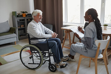 Full length side view portrait of white haired senior man with disability talking to smiling nurse in retirement home, copy space