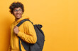 Student lifestyle. Studio sideways shot of young cheerful smiling Hindu male wearing yellow hoodie with big black bag standing on left isolated looking aside with blank space for your promotion
