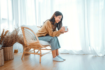 Happy asian woman relaxing drinking hot coffee or tea in holiday morning vacation on armchair at home, Cosy scene, Smiling pretty woman drinking hot tea in autumn winter. copy space.