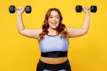 Young smiling strong happy chubby overweight plus size big fat fit woman wear blue top warm up training hold dumbbells wink isolated on plain yellow background studio home gym. Workout sport concept.
