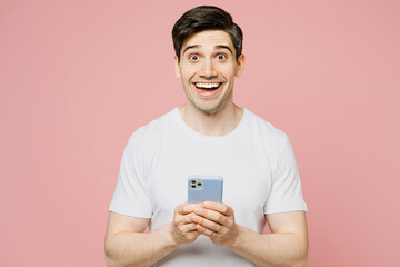 Wall Mural - Young surprised caucasian man wearing white t-shirt casual clothes hold in hand using mobile cell phone in blue case isolated on plain pastel light pink background studio portrait. Lifestyle concept.