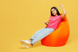 Full body young happy Indian woman wear pink t-shirt casual clothes sit in bag chair hold use mobile cell phone do winner gesture isolated on plain yellow background studio portrait Lifestyle concept