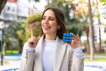Young pretty woman holding a three dimensional puzzle cube at outdoors intending to realizes the solution while lifting a finger up