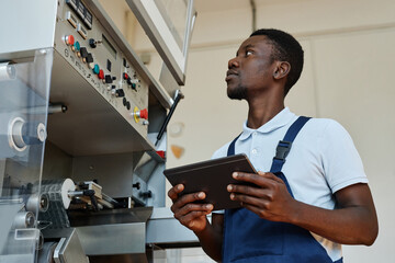 Wall Mural - Side view of African American factory worker holding tablet while operating machine units and overseeing production, copy space