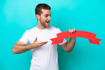 Wall Mural - Young caucasian man isolated on blue background holding an empty placard with surprised expression