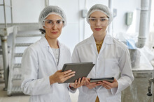 Waist Up Portrait Of Two Young Women Wearing Lab Coats Looking At Camera And Smiling While Using Digital Tablet In Workshop