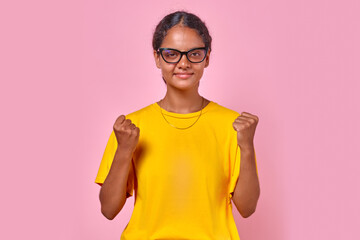 Young smiling ethnic Indian woman teenager makes victory gestures with both hands after learning about opportunity to go on trip with friends from high school or university stands on pink background.