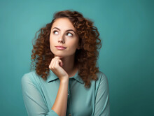 Headshot Portrait Of Thoughtful Pensive Young Ginger Woman With Curly Hair Holding Hand Under Chin Looking Upward Against Turquoise Studio Wall Background With Copy Space For Text Advertisement 