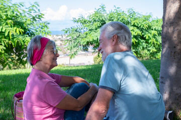 Smiling senior couple sitting in the park having relaxed moments together, retired grey-haired man and woman in the meadow looking each other and talking