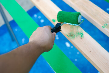 Hand using 6 inches paint roller for first coat of latex paint on 2 by 4 wooden boards on sawhorse with blue tarp cover paint drops outdoor in Dallas, Texas, USA