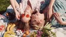 Two Young Beautiful Smiling Hipster Female In Trendy Summer Sundress And Hats. Carefree Women Making Picnic Outside. Positive Models Sitting On Plaid On Grass, Hugging, Eating Fruits. Top View