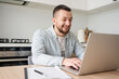© Julija - Portrait of young smiling man using laptop sitting at desk, writing in notebook. Cheerful guy browsing internet, watching webinar studying online, looking at pc screen at home.