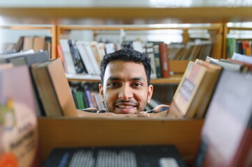 Poster - Portrait of cheerful male international Indian student with backpack, learning accessories standing near bookshelves at university library or book store during break between lessons. Education concept