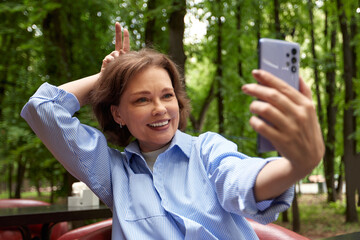 Wall Mural - Attractive smiling senior woman making selfie. Mature caucasian lady uses smartphone. Middle aged brunette female in blue shirt enjoying life outside in a park. Active lifestyle for the elderly.