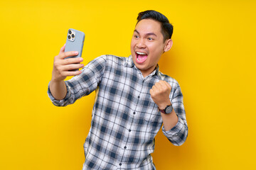 Wall Mural - Excited young Asian man wearing a white checkered shirt looking at a smartphone screen and celebrating success, reacting to online news isolated on yellow background. People Lifestyle Concept