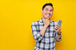 © Bangun Stock Photo - Smiling young Asian man wearing a white checkered shirt touching his chin and holding a smartphone, thinking about something isolated on yellow background. People Lifestyle Concept