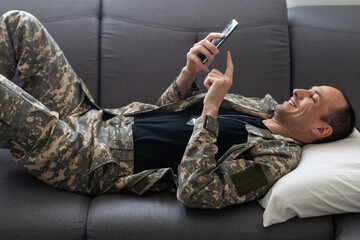 Horizontal shot of Caucasian military man wearing camouflage uniform sitting on sofa, soldier holding cell phone, looking at display, watching video or having online conversation.