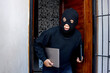 Sneaky and scared intruder wearing black balaclava mask stealing laptop from house. House intruder, housebreaking and burglary concept