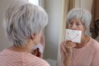 Sad senior woman holding a drawing with a smile in front of the mirror 