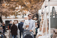 Father Walking Carrying His Infant Baby Boy Child And Pushing Stroller In Crowd Of People Wisiting Sunday Flea Market In Malaga, Spain