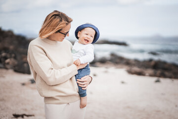 Wall Mural - Mother enjoying winter vacations holding and playing with his infant baby boy son on volcanic sandy beach on Lanzarote island, Spain. Family travel and vacations concept.