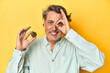 © Asier - Man holding a physical bitcoin, yellow backdrop excited keeping ok gesture on eye.