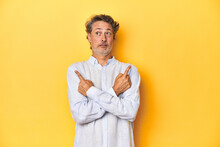 Middle-aged Man Posing On A Yellow Backdrop Points Sideways, Is Trying To Choose Between Two Options.
