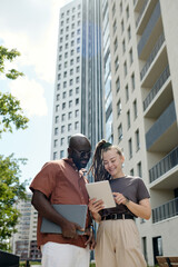 Fototapete - Happy young businesswoman with tablet making slide show to African American male colleague while standing next to him in the street
