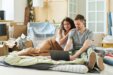 Wall Mural - Young affectionate couple sitting on checkered plaid on the floor of living room and looking at laptop screen while man typing on keyboard