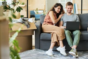 Wall Mural - Happy young couple relaxing on soft comfortable couch in living room and watching online video or movie while man holding laptop on his knees