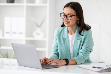 Wall Mural - Cheerful millennial caucasian lady in suit and glasses typing on laptop at workplace