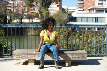 Young and beautiful black woman with afro hair dressed in jeans and yellow shirt sitting on a wooden bench, the woman is happy. In the background the river guadalquivir of seville in spain.