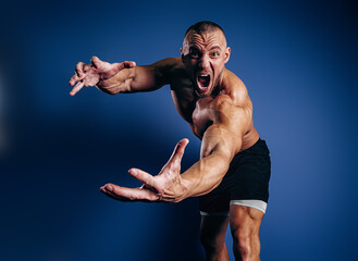 Super emotional bodybuilder screaming and showing muscles on blue background. Fit man with muscular body