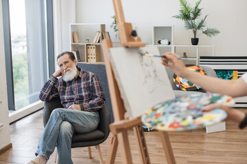 Selective focus of aged man in casual clothes sitting in chair behind wooden easel with painting in workshop. Grey-haired male in his 70s posing in studio interior during life drawing session.