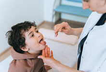 Female Doctor Checking Childs Throat Of Little Curly Boy Using Medical Wand For Examination At Hospital. Hispanic Toddler Shows Tongue To Nurse. Children Medical Care And Healthy Lifestyle.