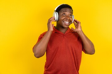 Joyful Young latin man wearing red t-shirt over yellow background sings song keeps hand near mouth as if microphone listens favorite playlist via headphones