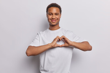 Sticker - Handsome dark skinned man bares his heart and confesses in love delicately forms heart gesture with fingers conveys romantic feelings passionate emotions wears casual t shirt isolated on white wall