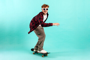 Wall Mural - young cheerful guy rides skateboard on blue isolated background, hipster skater in sunglasses