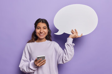 Waist up shot of pretty cheerful Iranian woman uses modern smartphone holds empty speech bubble suggests to write your text here smiles gladfully dressed in casual jumper isolated on purple background