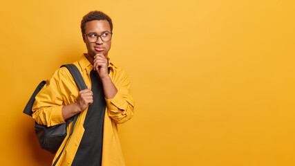 Wall Mural - Horizontal shot of thoughtful millennial guy keeps hand on chin concentrated aside pensively considers something wears spectacles and shirt carries rucksack isolated over vivid yellow background