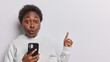 © wayhome.studio  - Oh no check this. Impressed curly haired woman with dark skin points index finger on blank space for your advertising content poses with smartphone dressed casually isolated over white background