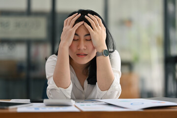 Portrait image of Young Asian businesswoman bowing and holding her head stressed expression with financial documents on working table.