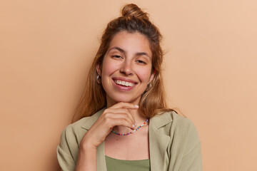 Poster - Portrait of cheerful long haired young woman touches chin gently exuding joy and contentment has happy expression enjoys delightful weekend moment wears shirt isolated over brown background.