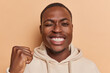 Cheerful black man with white teeth confidently makes winner gesture embodying sincere emotions winks eye stands happy anticipates for something awesome wears casual hoodie isolated over brown wall