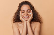 © wayhome.studio  - Horizontal shot of curly haired Brazilian woman applies facial cream for nurturing her complexion keeps eyes closed smiles boadly uses cosmetic product to nourish skin isolated over brown background