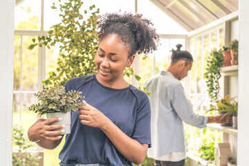 African American wife planting trees as a hobby and water the nursery and Asian husband decorating trees in the back of the green house in the backyard.