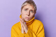 Headshot of blonde short haired young woman keeps hands in praying gesture with hope asking help and support has pity expression wears yellow jumper with collar isolated over purple background