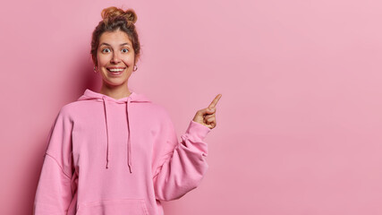 Wall Mural - Waist up shot of cheerful young European woman has surprised glad expresssion points index finger at empty space shows advertisement dressed in casual sweatshirt isolated over pink background