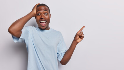 Wall Mural - Waist up shot of impressed dark skinned man keeps hand on head exclaims loudly reacts to unexpected offer points index finger at upper right corner dressed in blue t shirt isolated on white background