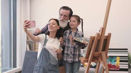 Wall Mural - Lovely family of three generations making selfie on personal smartphone while painting picture on wooden easel. Mother, daughter and granddad having fun together during weekend time in spacious flat.